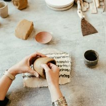 Osoba formuje naczynie z gliny na stole roboczym, na którym leżą różne narzędzia i kawałki gliny.