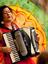 Kobieta o ciemnych kręconych włosach, ubrana w czerwoną sukienkę, gra na akordeonie i śpiewa. W tle widoczny jest kolorowy, dekoracyjny wzór w ciepłych odcieniach zieleni, żółci i pomarańczy.