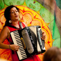 Kobieta o ciemnych kręconych włosach, ubrana w czerwoną sukienkę, gra na akordeonie i śpiewa. W tle widoczny jest kolorowy, dekoracyjny wzór w ciepłych odcieniach zieleni, żółci i pomarańczy.