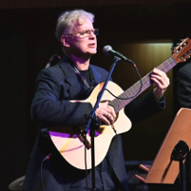 Zdjęcie mężczyzny na scenie, na której gra na gitarze i śpiewa. Przed nim stoi mikrofon na statywie.