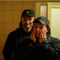 Dwóch mężczyzn w ciemnych ubraniach i czapkach z daszkiem stoi w słabo oświetlonym pomieszczeniu. Mężczyzna z przodu trzyma dłonie na policzkach, wyglądając na zmęczonego lub zrezygnowanego, podczas gdy drugi, stojący za nim się uśmiecha..