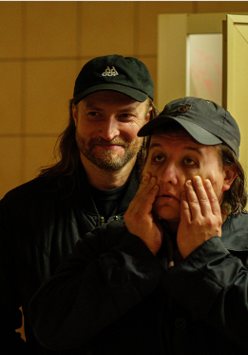 Dwóch mężczyzn w ciemnych ubraniach i czapkach z daszkiem stoi w słabo oświetlonym pomieszczeniu. Mężczyzna z przodu trzyma dłonie na policzkach, wyglądając na zmęczonego lub zrezygnowanego, podczas gdy drugi, stojący za nim się uśmiecha..