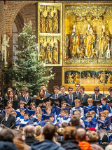 Chór dziecięco-młodzieżowy i dorośli śpiewacy stoją w prezbiterium zabytkowego kościoła, trzymając nuty i śpiewając przed zgromadzoną publicznością. Za nimi bogato zdobiony złoty ołtarz oraz choinki bożonarodzeniowe.