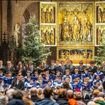 Chór dziecięco-młodzieżowy i dorośli śpiewacy stoją w prezbiterium zabytkowego kościoła, trzymając nuty i śpiewając przed zgromadzoną publicznością. Za nimi bogato zdobiony złoty ołtarz oraz choinki bożonarodzeniowe.