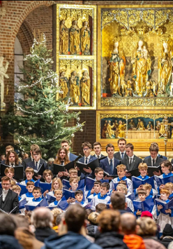Chór dziecięco-młodzieżowy i dorośli śpiewacy stoją w prezbiterium zabytkowego kościoła, trzymając nuty i śpiewając przed zgromadzoną publicznością. Za nimi bogato zdobiony złoty ołtarz oraz choinki bożonarodzeniowe.