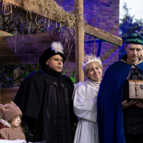 Plenerowa scena jasełek. Widać postacie w historycznych strojach stoją przy drewnianej stajence z sianem. Maria i Józef z dzieckiem w żłóbku, aniołowie oraz trzej królowie, jeden trzyma ozdobną szkatułkę i gestem zwraca się do publiczności.