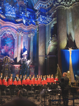 Na zdjęciu chór śpiewający w poznańskiej farze, widać chórzystów, podświetlony ołtarz i dyrygenta.