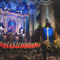 Na zdjęciu chór śpiewający w poznańskiej farze, widać chórzystów, podświetlony ołtarz i dyrygenta.
