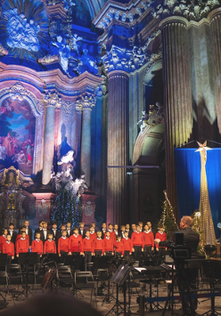 Na zdjęciu chór śpiewający w poznańskiej farze, widać chórzystów, podświetlony ołtarz i dyrygenta.