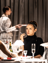 Grupa osób siedzących przy stole, obsługiwanych przez kelnerkę, w tle widac kelnera. Kobieta na środku zdjęcia trzyma w ręce papierosa. Na stole widać kieliszki i butelkę.