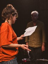 Kobieta oraz mężczyzna w półmroku na scenie. Kobieta jest zwrócona bokiem i w lewej ręce trzyma kartkę. Mężczyzna patrzy na kobietę na wprost.