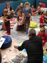 Grupa dzieci i dorosłych siedzi na podłodze w sali warsztatowej, wspólnie malując kolorowe obrazy na kartkach rozłożonych na folii ochronnej. Wokół widać pufy, farby i rysunki w trakcie tworzenia.