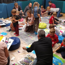 Grupa dzieci i dorosłych siedzi na podłodze w sali warsztatowej, wspólnie malując kolorowe obrazy na kartkach rozłożonych na folii ochronnej. Wokół widać pufy, farby i rysunki w trakcie tworzenia.