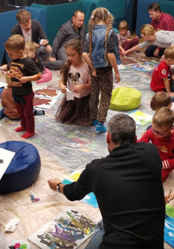 Grupa dzieci i dorosłych siedzi na podłodze w sali warsztatowej, wspólnie malując kolorowe obrazy na kartkach rozłożonych na folii ochronnej. Wokół widać pufy, farby i rysunki w trakcie tworzenia.