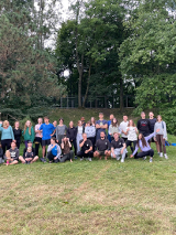 Gruba ludzi ubranych w kolorowych strojach stojących i siedzących na trawie. W tle widać drzewa.