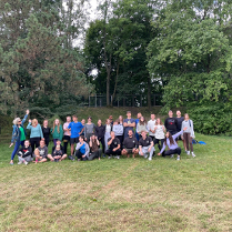 Gruba ludzi ubranych w kolorowych strojach stojących i siedzących na trawie. W tle widać drzewa.