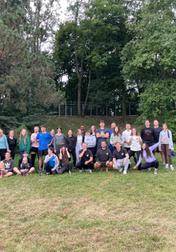 Gruba ludzi ubranych w kolorowych strojach stojących i siedzących na trawie. W tle widać drzewa.