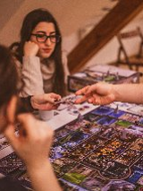 Grupa osób skupiona wokół stołu podczas rozgrywki w rozbudowaną grę planszową. Na pierwszym planie widoczne plecy i profil ciemnowłosej kobiety oraz dłoń gracza sięgająca po kartę na kolorowej, szczegółowej planszy. W centralnej części dwie młode kobiety z uwagą obserwują przebieg gry. Jedna z nich, w okularach i beżowym swetrze opiera podbródek na dłoni. Tło tworzy przytulne, domowe wnętrze z drewnianymi schodami i krzesłami w ciepłym, przygaszonym świetle.