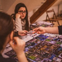 Grupa osób skupiona wokół stołu podczas rozgrywki w rozbudowaną grę planszową. Na pierwszym planie widoczne plecy i profil ciemnowłosej kobiety oraz dłoń gracza sięgająca po kartę na kolorowej, szczegółowej planszy. W centralnej części dwie młode kobiety z uwagą obserwują przebieg gry. Jedna z nich, w okularach i beżowym swetrze opiera podbródek na dłoni. Tło tworzy przytulne, domowe wnętrze z drewnianymi schodami i krzesłami w ciepłym, przygaszonym świetle.