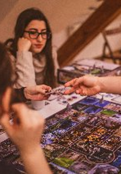 Grupa osób skupiona wokół stołu podczas rozgrywki w rozbudowaną grę planszową. Na pierwszym planie widoczne plecy i profil ciemnowłosej kobiety oraz dłoń gracza sięgająca po kartę na kolorowej, szczegółowej planszy. W centralnej części dwie młode kobiety z uwagą obserwują przebieg gry. Jedna z nich, w okularach i beżowym swetrze opiera podbródek na dłoni. Tło tworzy przytulne, domowe wnętrze z drewnianymi schodami i krzesłami w ciepłym, przygaszonym świetle.