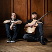 Dwóch mężczyzn siedzących na podłodze pod brązową, drewnianą ścianą z dekoracyjnymi kasetonami. Po lewej stronie mężczyzna z dłuższymi ciemnymi włosami, ubrany w szarą kamizelkę, koszulę i ciemne spodnie, z dłońmi opartymi na kolanach. Po prawej stronie mężczyzna w czapce z daszkiem, okularach i czarnym ubraniu, trzymający na kolanach jasną gitarę akustyczną. Obaj patrzą bezpośrednio w obiektyw z poważnym wyrazem twarzy.