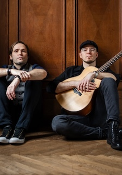 Dwóch mężczyzn siedzących na podłodze pod brązową, drewnianą ścianą z dekoracyjnymi kasetonami. Po lewej stronie mężczyzna z dłuższymi ciemnymi włosami, ubrany w szarą kamizelkę, koszulę i ciemne spodnie, z dłońmi opartymi na kolanach. Po prawej stronie mężczyzna w czapce z daszkiem, okularach i czarnym ubraniu, trzymający na kolanach jasną gitarę akustyczną. Obaj patrzą bezpośrednio w obiektyw z poważnym wyrazem twarzy.