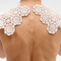 Artystyczne ujęcie przedstawiające nagie plecy i ramiona osoby, na których ułożona jest biała, koronkowa serweta wykonana z połączonych okrągłych elementów o kwiatowym wzorze. Zdjęcie wykonane na jednolitym białym tle.