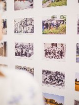 Na białej ścianie zawieszonych jest w równych rzędach kilkanaście małych, poziomych zdjęć prezentujących różne sceny: od ujęć architektury i budynków, przez portrety grupowe, aż po fotografie przyrody. Na pierwszym planie, w znacznym rozmyciu (nieostrości), widać fragmenty głowy i ramiona dwóch osób, które przyglądają się ekspozycji. Jedna z osób ma na sobie jasną bluzkę, a druga niebieską w drobny wzór.