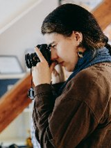 Kobieta w warkoczu, w brązowej kurtce z niebieskim szalem, trzyma aparat przy twarzy i robi zdjęcie. W tle widać drewniane belki i półki z przedmiotami.