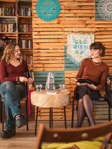Dwie kobiety siedzą we wnętrzu biblioteki lub czytelni, rozmawiają przy okrągłym stole z dzbankiem wody. W tle widać półki z książkami i kolorowe dekoracje na drewnianej ścianie. Na pierwszym planie widać rozmazane krzesło i tył głowy osoby siedzącej od strony publiczności.