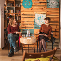Dwie kobiety siedzą we wnętrzu biblioteki lub czytelni, rozmawiają przy okrągłym stole z dzbankiem wody. W tle widać półki z książkami i kolorowe dekoracje na drewnianej ścianie. Na pierwszym planie widać rozmazane krzesło i tył głowy osoby siedzącej od strony publiczności.
