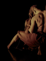 Zdjęcie przedstawiające mężczyznę z odsłoniętymi plecami i ramionami, częściowo okrytego tkaniną. Postać jest ukazana w półmroku na czarnym tle, z widocznym światłem podkreślającym mięśnie mężczyzny i fakturę materiału.