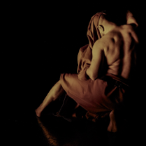 Zdjęcie przedstawiające mężczyznę z odsłoniętymi plecami i ramionami, częściowo okrytego tkaniną. Postać jest ukazana w półmroku na czarnym tle, z widocznym światłem podkreślającym mięśnie mężczyzny i fakturę materiału.
