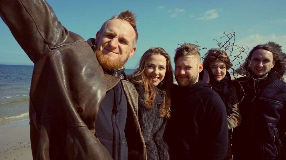 Zespół pozuje nad morzem, dwie kobiety i trzech mężczyzn. Stoją blisko siebie, przytulając się i uśmiechając.