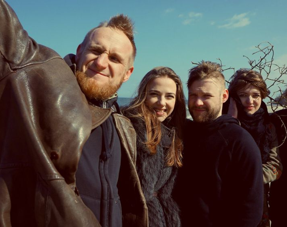Zespół pozuje nad morzem, dwie kobiety i trzech mężczyzn. Stoją blisko siebie, przytulając się i uśmiechając.
