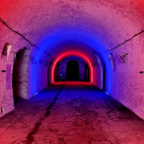 Zdjęcie przedstawia długi ceglany tunel oświetlony kolorowymi światłami. Lewa ściana jest oświetlona czerwonym światłem, a prawa ściana jest oświetlona niebieskim światłem. Tunel jest pusty, a jedynym źródłem światła są światła kolorowe. Zdjęcie zostało zrobione z centrum tunelu, patrząc w stronę wyjścia.
