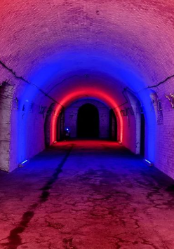 Zdjęcie przedstawia długi ceglany tunel oświetlony kolorowymi światłami. Lewa ściana jest oświetlona czerwonym światłem, a prawa ściana jest oświetlona niebieskim światłem. Tunel jest pusty, a jedynym źródłem światła są światła kolorowe. Zdjęcie zostało zrobione z centrum tunelu, patrząc w stronę wyjścia.