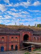 Fort zbudowany z czerwonych cegieł z licznymi okiennicami i głównym wejściem, do którego prowadzi most. Nad fortem na zielenii stoi sztandar z polską flagą. W tle zielone drzewa i niebieskie niebo z chmurami.