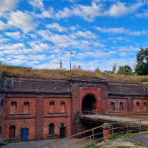 Fort zbudowany z czerwonych cegieł z licznymi okiennicami i głównym wejściem, do którego prowadzi most. Nad fortem na zielenii stoi sztandar z polską flagą. W tle zielone drzewa i niebieskie niebo z chmurami.