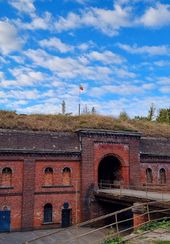 Fort zbudowany z czerwonych cegieł z licznymi okiennicami i głównym wejściem, do którego prowadzi most. Nad fortem na zielenii stoi sztandar z polską flagą. W tle zielone drzewa i niebieskie niebo z chmurami.