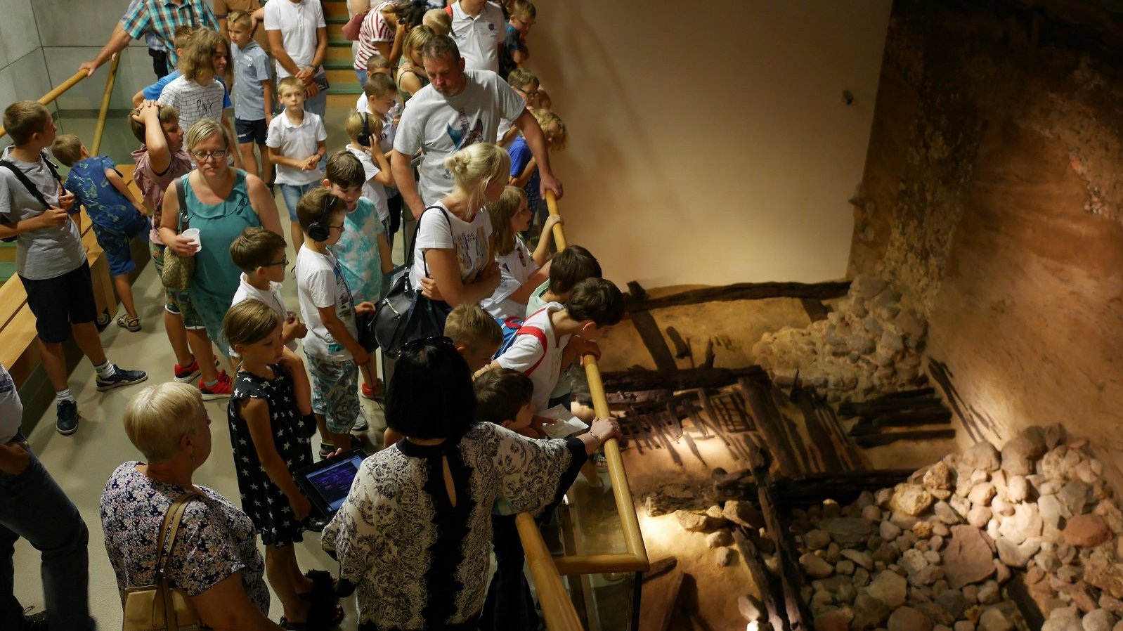 Duża grupa zwiedzających, w różnym wieku, zwiedza ekspozycje i spogląda na zachowane relikty wału obronnego Ostrowa Tumskiego z czasów piastowskich.