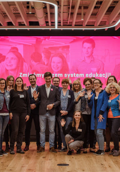 Zdjęcie grupowe. Grupa stojacych ludzi na scenie. W tle rózowy ekran z młodzieżą.
