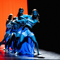 A few women, lit by blue light, in a row in dance poses. Black and orange background.