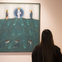 A girl visiting the exhibition is looking at one of the icons - a large one, on which an image of a biblical scene has been painted. The dominant colors are blue and green.