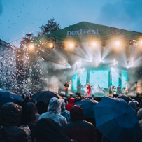 A lit stage with a band and a singer, and a crowd of people with umbrellas in front of it