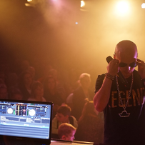 A man in a black T-shirt with a slogan, a chain around his neck and sunglasses, on stage, his back to the audience. On the left side, a screen displays blue graphics. An audience in a background.