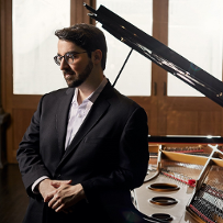 A man in a shirt and jacket stands at the grand piano.