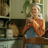A smiling girl stands with a mug in her hands, leaning against the kitchen counter. There are kitchen cabinets and a small window behind her.
