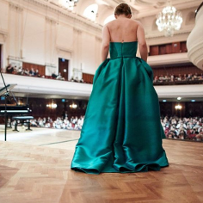 A woman in an elegant, green dress on a stage, standing with her back to the camera. An audience in a background.