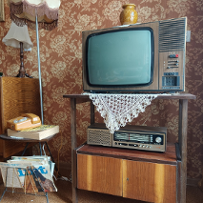 An old television set standing on a small table, under it old-fashioned radio. On the left a lamp and a small stand on which a dial telephone and a phone book are lying. Patterned white and brown wallpaper as a background.